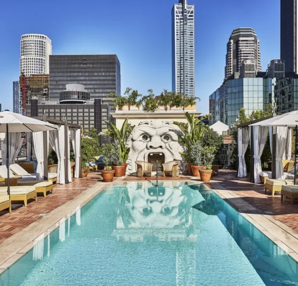 Luxurious rooftop pool deck with turquoise swimming pool surrounded by white cabanas and yellow wicker lounge chairs. A large sculptural face with an open mouth serves as a decorative water feature against the back wall, surrounded by potted plants. The downtown Los Angeles skyline with modern high-rise buildings rises in the background under a clear blue sky