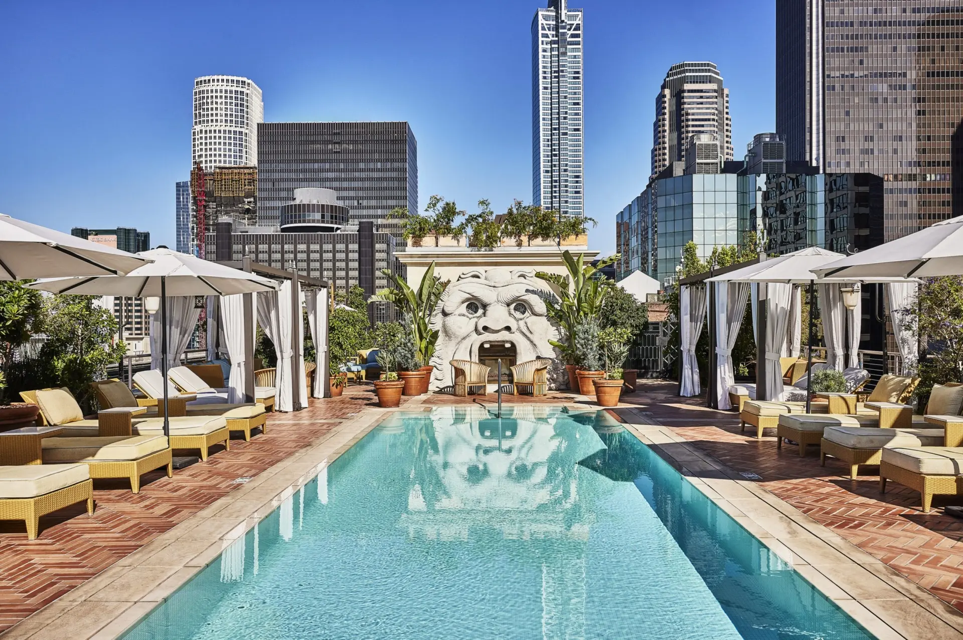 Luxurious rooftop pool deck with turquoise swimming pool surrounded by white cabanas and yellow wicker lounge chairs. A large sculptural face with an open mouth serves as a decorative water feature against the back wall, surrounded by potted plants. The downtown Los Angeles skyline with modern high-rise buildings rises in the background under a clear blue sky