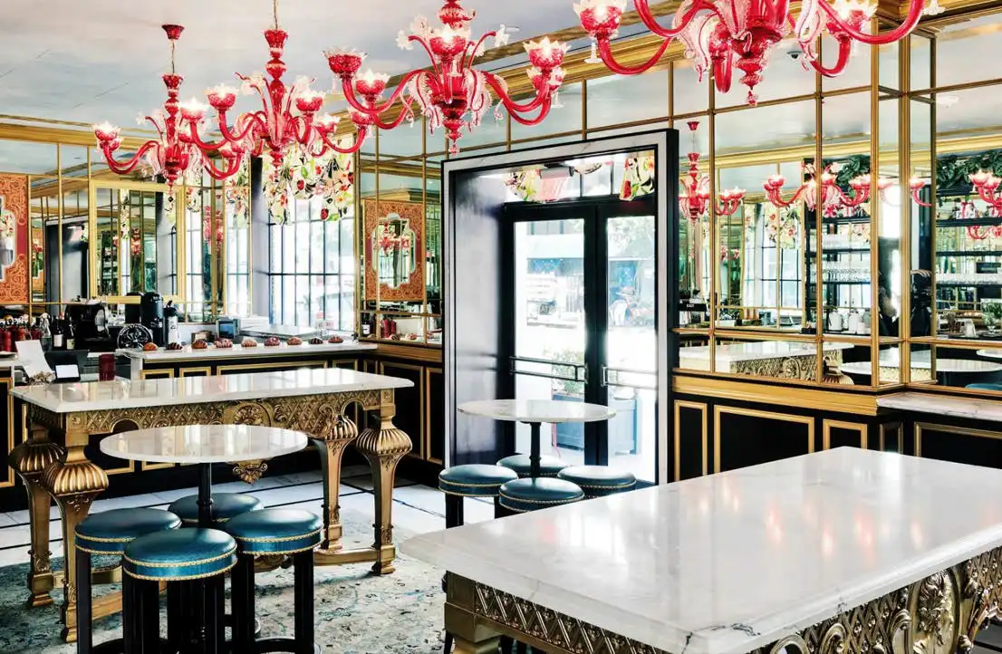 Elegant red glass chandeliers hang over marble tables and teal stools in the vibrant Cafe Ora dining area.