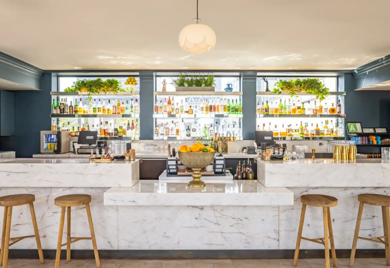 A bright marble bar with wooden stools and greenery at Bar Clara, a premier rooftop bar in Los Angeles CA.