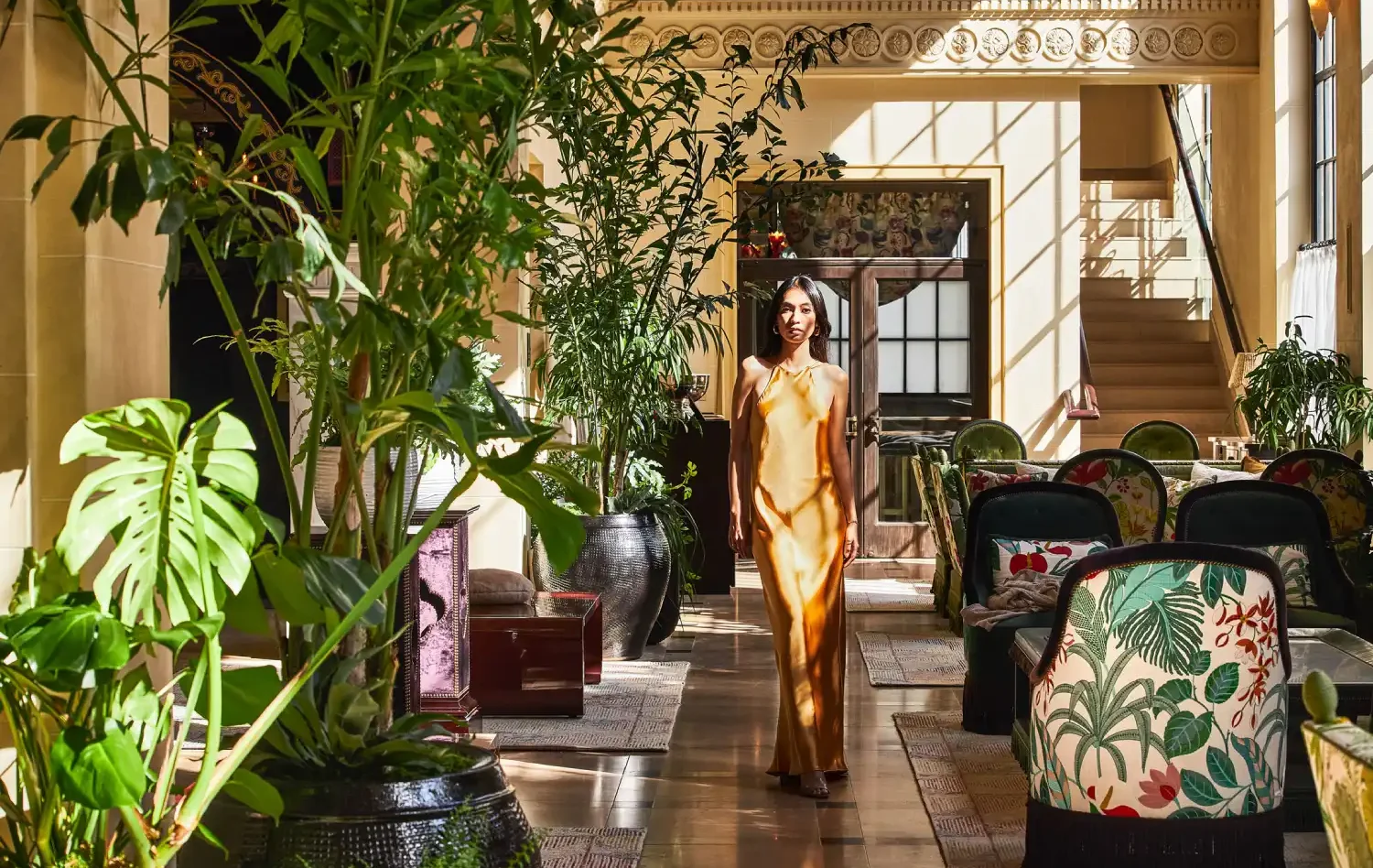 A woman in a gold dress walks through a sunlit lobby filled with lush plants and floral armchairs.