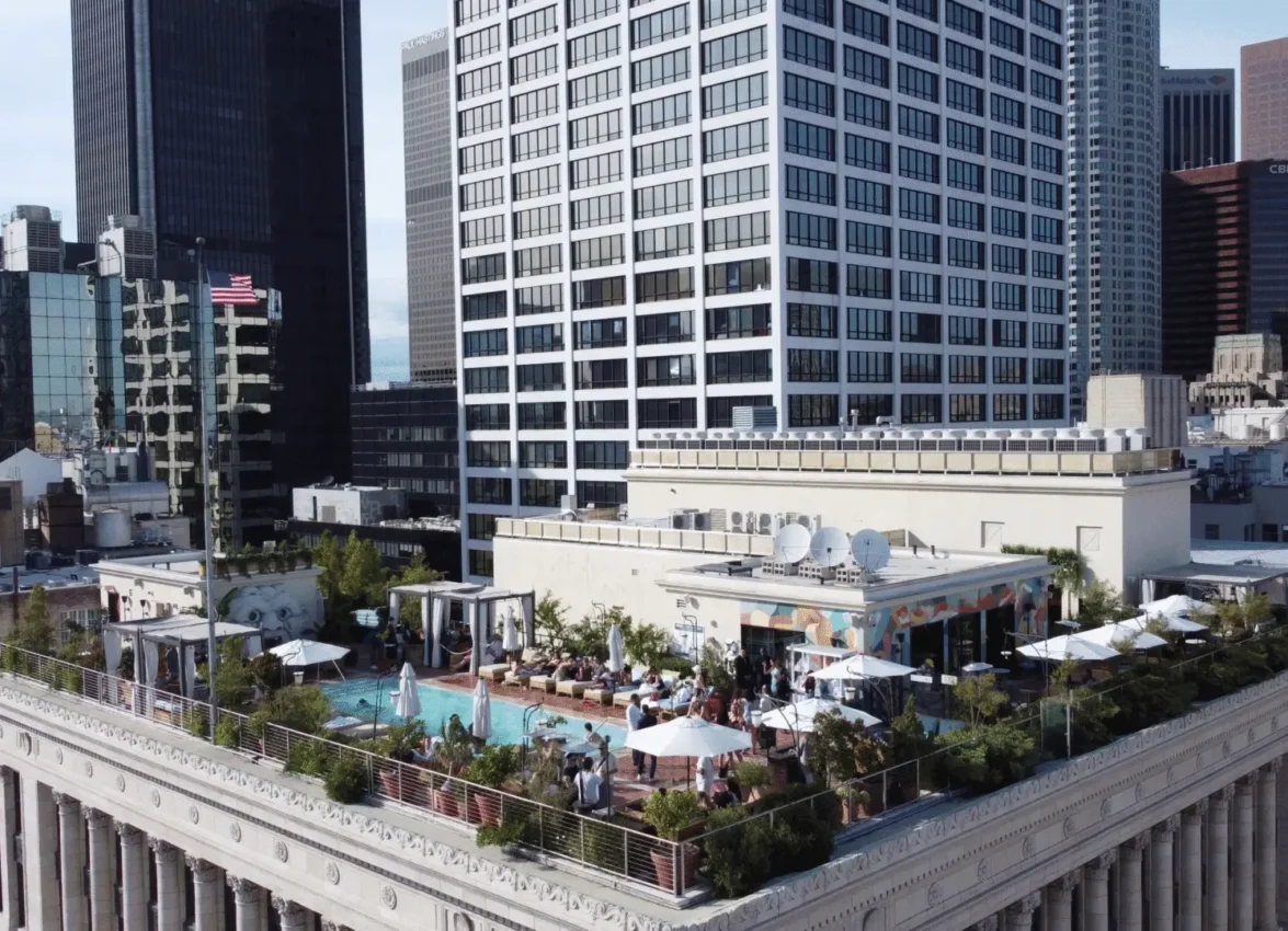 Aerial view of a bustling rooftop venue with swimming pool, dining areas, and white umbrellas on top of a classical building with columned architecture. The rooftop space features a turquoise pool, multiple seating areas with guests, and colorful murals on building walls. The venue is surrounded by downtown high-rise buildings and skyscrapers, creating an urban oasis setting.