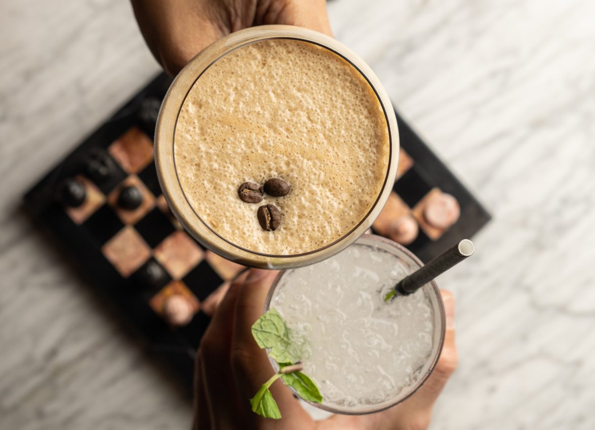 Two people toast an espresso martini and mint cocktail over a marble table and chessboard at Bar Clara in downtown LA.