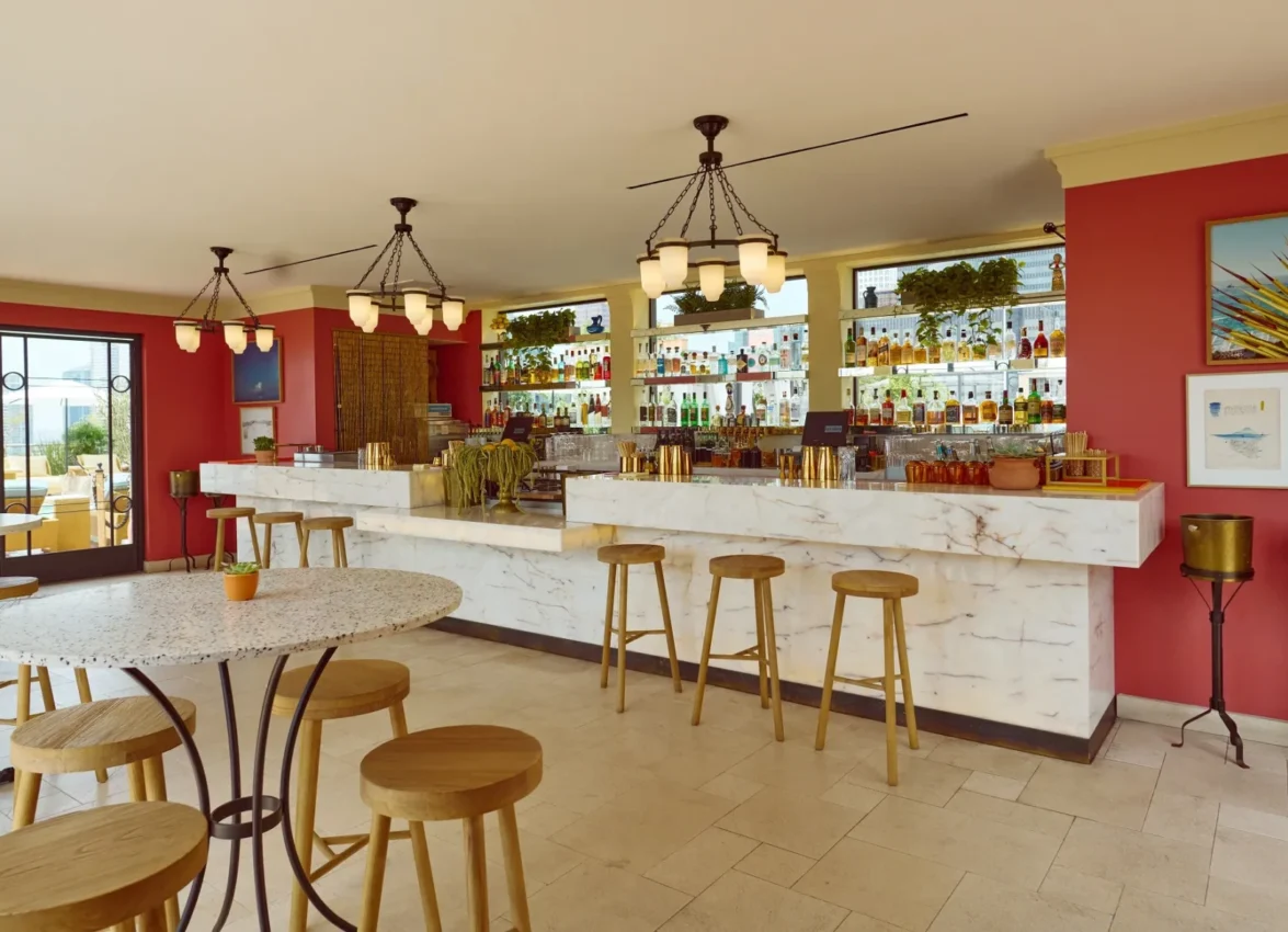 Modern rooftop bar interior featuring a white marble bar with wooden bar stools, coral-red accent walls, and an extensive backlit liquor display. The space includes round marble-top tables with wooden stools, industrial-style pendant lighting with frosted glass shades, and large windows offering city skyline views. Gold planters and tropical plants add warmth to the contemporary design.
