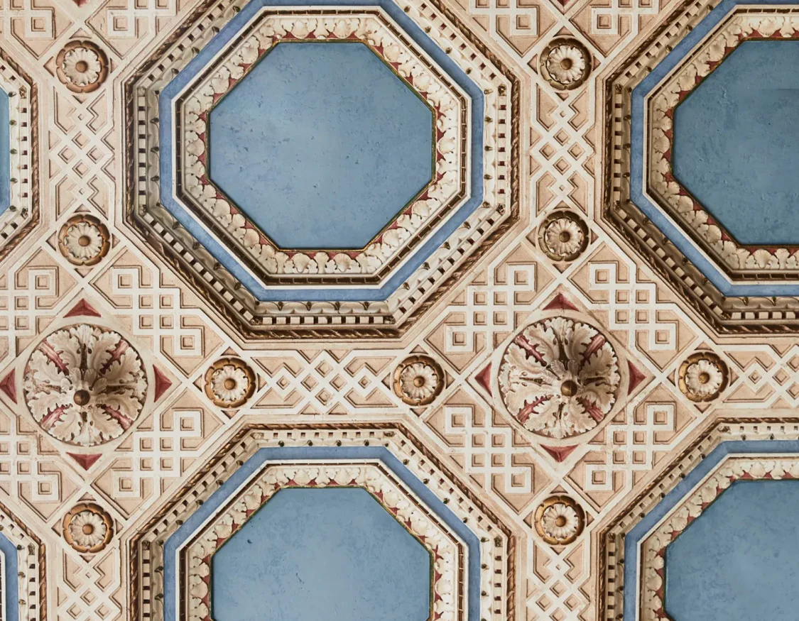 An ornate coffered ceiling with blue octagonal panels and intricate carvings highlights the luxury of Hotel Per La.