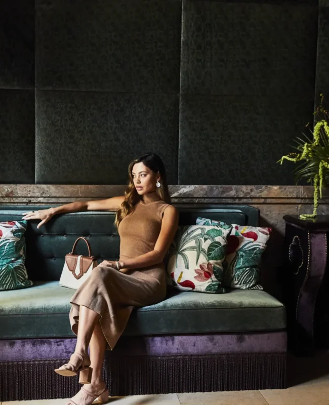 A woman walks through the sunlit, plant-filled lobby of Hotel Per La featuring ornate ceilings and floral seating.
